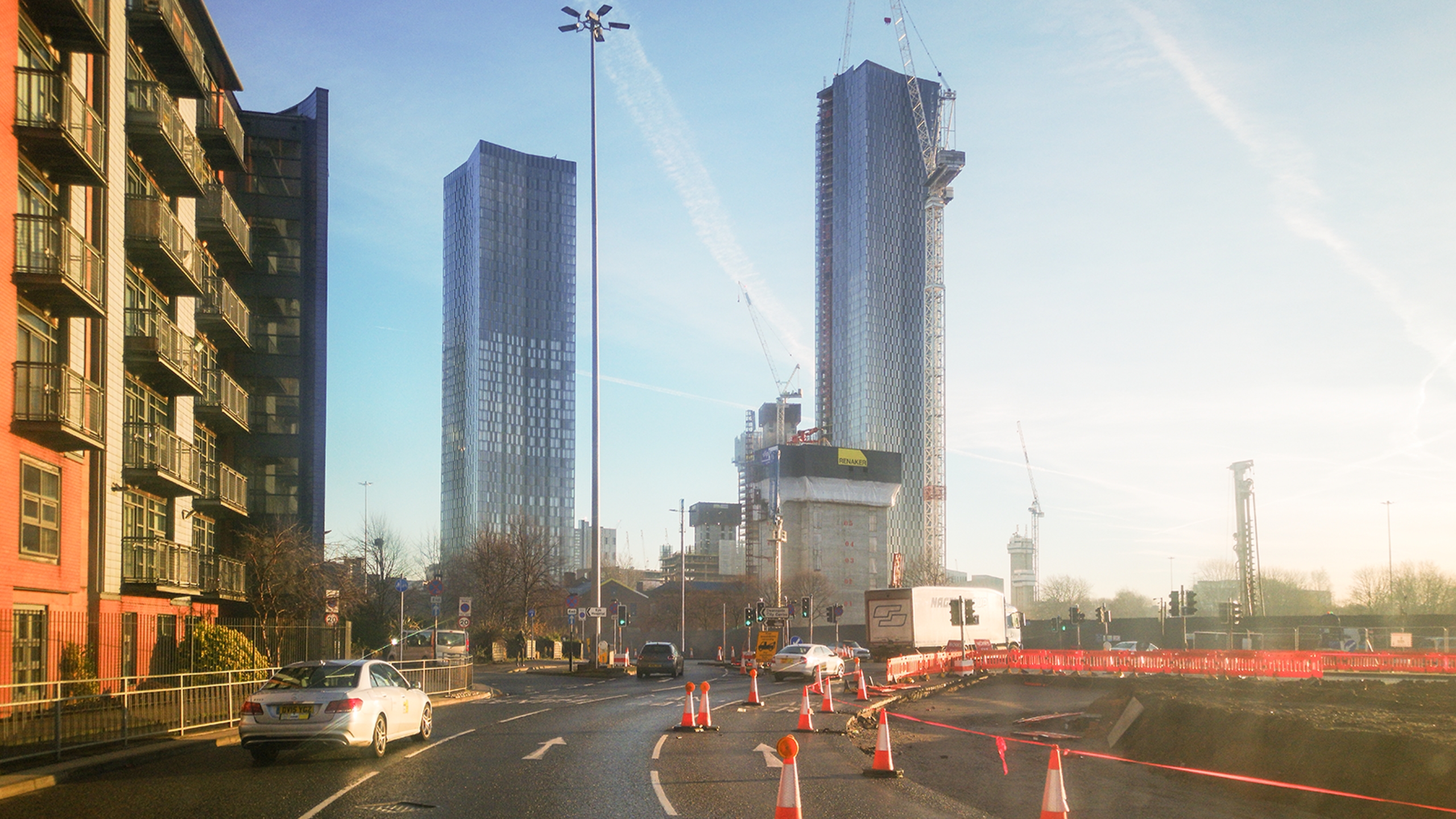 An image of central Manchester, shot from the Mancunian Way. The road is dotted with traffic cones; ugly new-build flats loom to the left, and in the background, we see the glass-and-steel towers of Deansgate Square, surrounded by cranes.