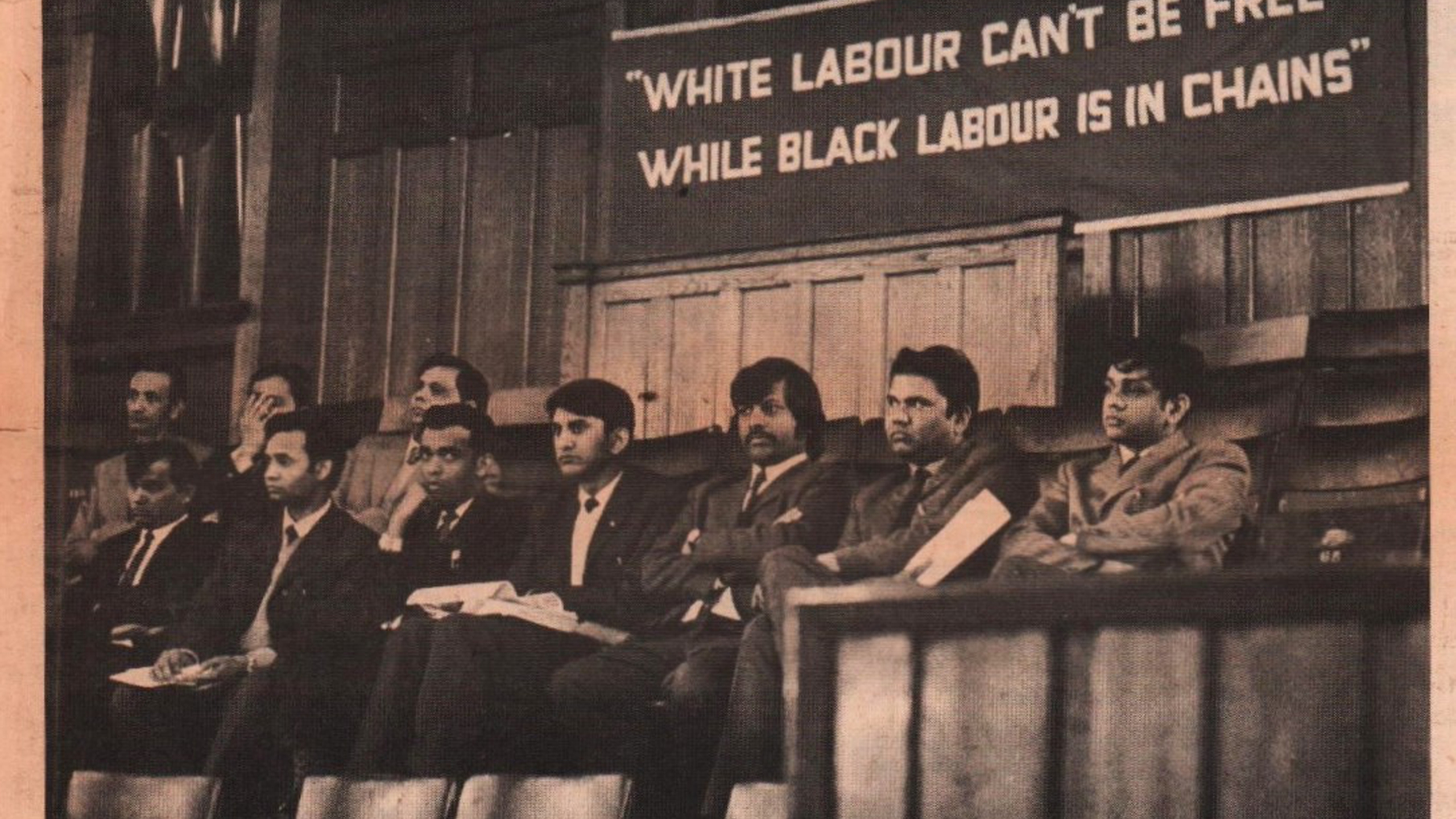 Photograph with banner in the background displaying the words "white labour can't be free while black labour is in chains". In the foreground, a group of racialised men wearing suits.